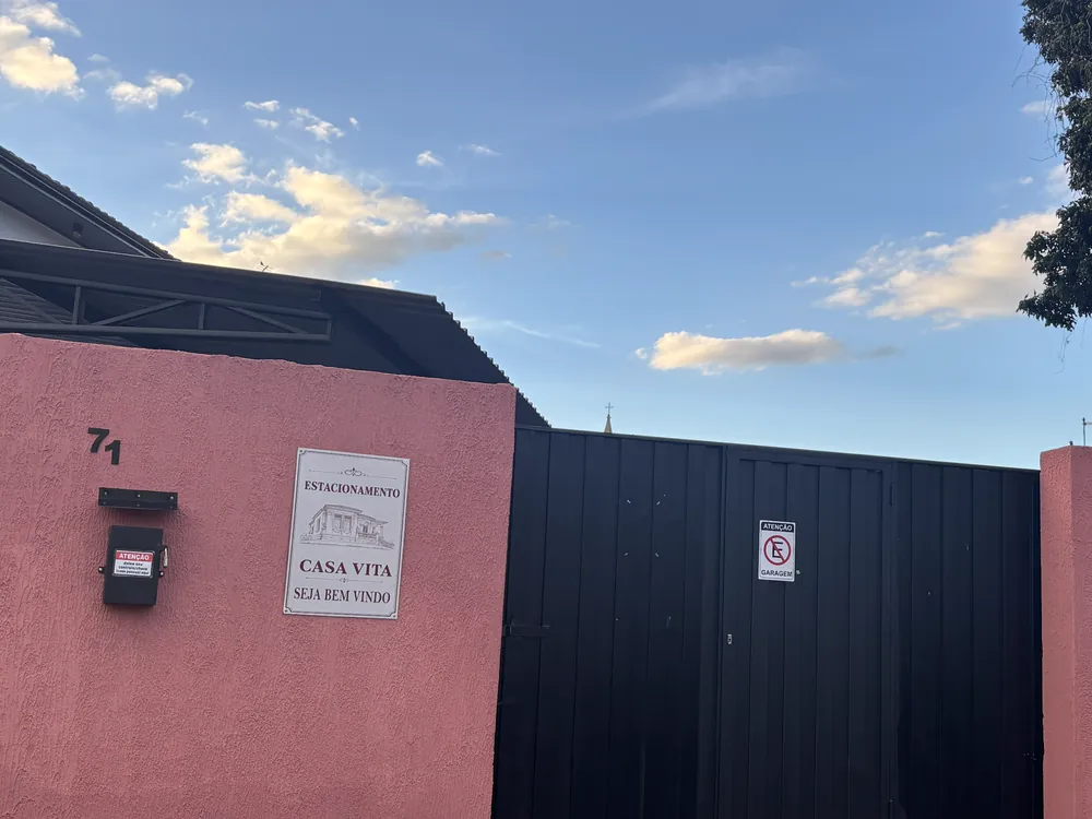 Portão preto do estacionamento ao lado da parede rosa com a placa da Casa Vita e o número 71